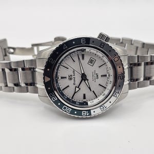 May include: A Grand Seiko Hi-Beat 36000 GMT watch with a silver textured dial, silver hands, and a date window. The watch features a black and silver rotating bezel with 24-hour markings and a brushed stainless steel link bracelet.