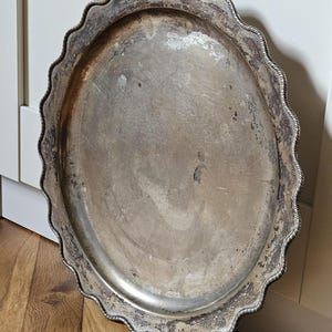 Könnte beinhalten: Ein ovales silberfarbenes Serviertablett mit gewelltem Rand und einer seilartigen Umrandung. Die Oberfläche des Tabletts weist Anzeichen von natürlicher Oxidation und Abnutzung auf, mit einem angelaufenen Finish in grauer Metalloptik.