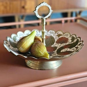 Könnte beinhalten: Eine Servierschale aus Messing mit gewelltem Rand und zentralem Griff, verziert mit einem komplexen Blumenmuster in grüner, roter und weißer Emaille. Zwei frische Feigen liegen in der Schale, die auf einem runden Messingsockel steht.