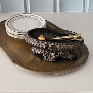 Peut inclure: Une pile d'assiettes blanches à bords floraux dorés et bleus, un bol ovale en métal sombre avec des détails en relief sur quatre pieds, et un plateau de service en métal doré. Une pince à sucre dorée repose dans le bol.