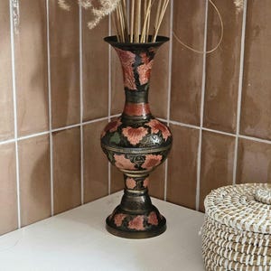 May include: A tall, ornate vase with a black base and a flared top. The vase features a floral design in shades of red and pink against a black background. Dried grasses are placed inside the vase. A woven basket is next to the vase.