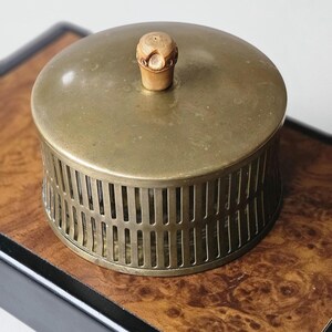 Pode incluir: Um recipiente redondo de latão antigo com tampa e um botão decorativo de madeira. O recipiente tem um design ranhurado nas laterais. A tampa é de cor latão maciço. O recipiente está sobre uma superfície de madeira.