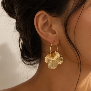 Op de afbeelding: Een gouden oorring met een gestructureerde, meerlaagse bloemhanger. De oorbel bestaat uit een dunne, ronde draadring waaraan een metalen bloem met opvallende, generfde bloemblaadjes hangt, wat zorgt voor een gelaagd en ruimtelijk effect.