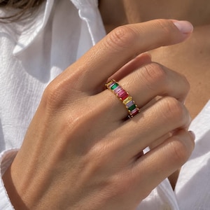 Può includere: Un anello a fascia color oro caratterizzato da una serie di pietre preziose rettangolari taglio baguette in un vibrante spettro arcobaleno, che include tonalità di verde, rosa, rosso, giallo e viola, disposte in fila continua.