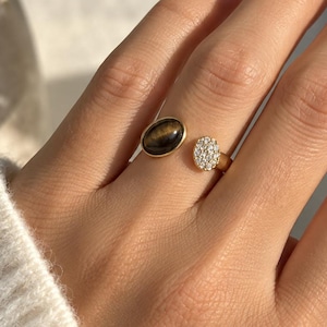 May include: A gold open-style ring featuring two distinct oval-shaped ends. One end holds a smooth, dark brown tiger's eye gemstone, while the other end is encrusted with a cluster of small, clear, sparkling white round-cut stones.