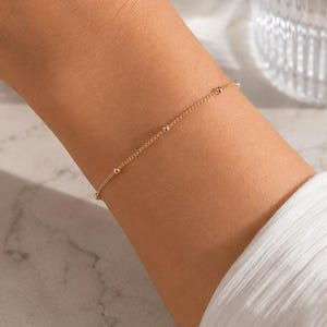 May include: A delicate gold chain bracelet featuring small, evenly spaced gold beads along the length of the chain. The bracelet is thin and minimalist in its construction, showcasing a simple link design with polished metallic spheres.