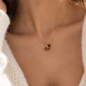 Pode incluir: Um colar de corrente dourada com um pequeno pingente em forma de romã com detalhes em pedras vermelhas e um berloque redondo vermelho. A delicada corrente de metal repousa sobre o pescoço, destacando o design detalhado da joia.