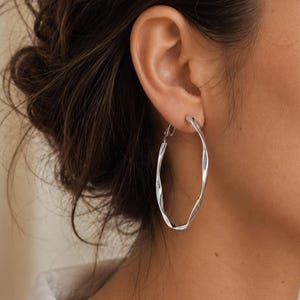 May include: A large silver-toned hoop earring with a twisted, spiraling metal texture. The earring features a circular design with a polished, reflective surface and a secure hinged closure mechanism visible at the top of the hoop.