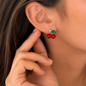 Puede incluir: Un pendiente de tipo botón con dos pequeñas cuentas esféricas de color rojo brillante con forma de cerezas, unidas a un fino tallo dorado con una hoja verde. El pendiente se sujeta al lóbulo con un pequeño cierre redondo de color dorado.
