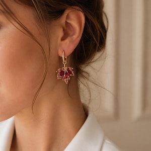 May include: A gold hoop earring featuring a dangling maple leaf charm. The hoop is accented with small clear stones, while the leaf charm is encrusted with deep red marquise-cut crystals and smaller red stones, creating a vibrant, detailed design.