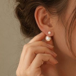 Peut inclure: Une boucle d'oreille à double face composée de deux perles sphériques blanches de tailles différentes. La plus petite perle se place sur le devant du lobe, tandis que la plus grande est positionnée derrière, créant un aspect superposé.