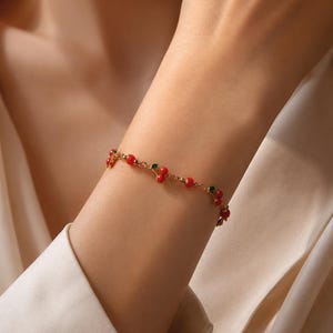 Peut inclure: Un bracelet à chaîne dorée orné de multiples petites breloques en forme de cerises rouges avec de minuscules feuilles vertes. La fine chaîne à maillons relie chaque motif de cerise, créant un motif répétitif de fruits le long du bijou.