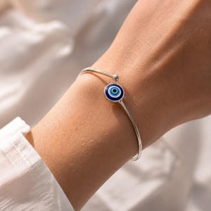 Könnte beinhalten: Ein silberfarbenes, offenes Armband mit einem runden Nazar-Amulett. Das Amulett zeigt konzentrische Kreise in Dunkelblau, Hellblau und Weiß mit einer schwarzen Pupille in der Mitte. Das Armband endet an beiden Öffnungen in kleinen silbernen Kugeln.