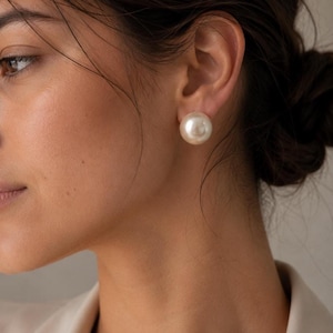Puede incluir: Un pendiente de botón con una perla grande, redonda y de color crema en el lóbulo de la oreja. La perla tiene una superficie lisa y brillante con un sutil lustre que refleja la luz uniformemente sobre su forma esférica, sujeta con un poste.