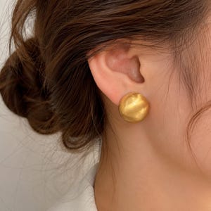 Può includere: Un orecchino a lobo circolare con finitura oro opaco e una texture superficiale leggermente bombata e irregolare. L'orecchino è di forma rotonda e presenta un aspetto metallico spazzolato e morbido che cattura la luce sulla superficie curva.