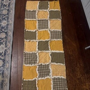 May include: A rectangular rag quilt runner featuring a patchwork design of alternating squares in mustard yellow, olive green, and dark green plaid patterns. The fabric squares are joined with exposed, frayed seams, creating a textured, layered appearance.