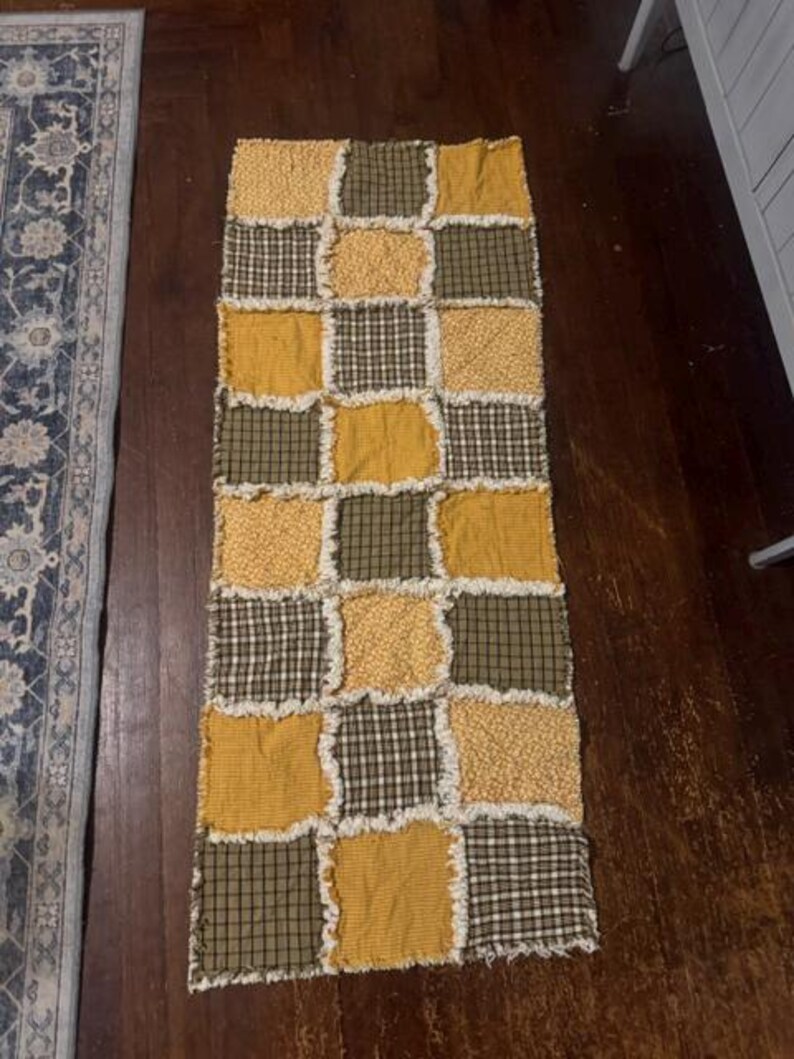 May include: A rectangular rag quilt runner featuring a patchwork design of alternating squares in mustard yellow, olive green, and dark green plaid patterns. The fabric squares are joined with exposed, frayed seams, creating a textured, layered appearance.