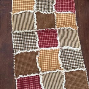 May include: A rectangular rag quilt table runner featuring a patchwork design of square fabric pieces in shades of red, tan, brown, and cream. The squares display various plaid and gingham patterns with exposed, frayed white seams between each block.