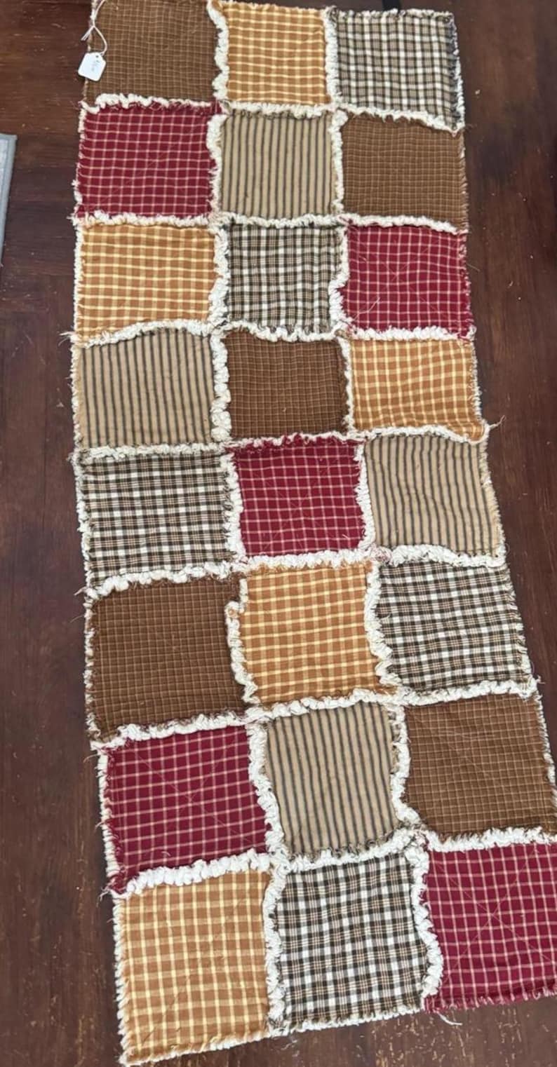 May include: A rectangular rag quilt table runner featuring a patchwork design of square fabric pieces in shades of red, tan, brown, and cream. The squares display various plaid and gingham patterns with exposed, frayed white seams between each block.