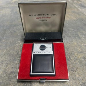 Puede incluir: Afeitadora Remington 300 Selectro vintage en su estuche original. La afeitadora es negra y plateada, con un agarre de cuero negro. El estuche es plateado con un interior de terciopelo rojo. El texto "Remington 300 Selectro Shaver" está impreso en el estuche.