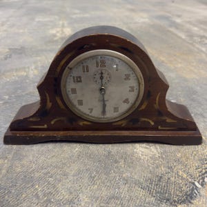 May include: An antique wooden clock with a round face and a dark brown finish. The clock face has black numerals and hands, with the brand name "Gilbert" visible. The clock has a decorative, arched top and a rectangular base.