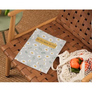 May include: A notebook with a daisy pattern and the text "MY FLOWER NOTES" rests on a brown leather chair. A white mesh bag filled with oranges sits nearby. The notebook has a light gray background with white daisies and green leaves.