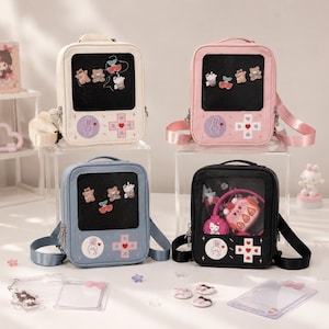 May include: Four rectangular backpacks in cream, pink, blue, and black, each featuring a clear front window, a top handle, and adjustable straps. The lower section of each bag displays a game controller design with a circular button and a cross-shaped button.