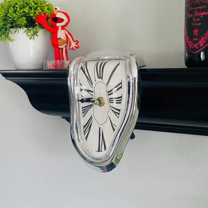 Op de afbeelding: Een zilverkleurige, smeltende klok met een witte wijzerplaat en zwarte Romeinse cijfers. De klok heeft een vervormd, surrealistisch ontwerp. De klok staat op een zwarte plank, met een kleine plant en een rood speelgoed links en een fles champagne rechts.