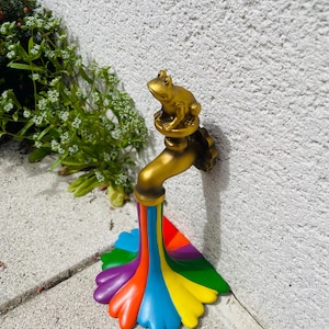 Puede incluir: Una escultura de grifo de jardín caprichosa con acabado dorado. Una pequeña rana dorada se sienta encima del grifo. El agua fluye en un vibrante arcoíris de colores, creando una pieza decorativa única y llamativa.