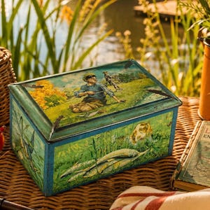 Op de afbeelding: Vintage metalen opbergdoos met een scharnierend deksel. De doos heeft een geschilderde scène van een jongen die aan het vissen is, met een hond en vissen. De doos heeft een blauw-groen kleurenschema, met een verweerde afwerking.
