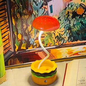 Peut inclure: Une lampe fantaisiste en forme de champignon avec un chapeau rouge et une tige blanche incurvée. La base est conçue pour ressembler à un hamburger, avec des couches jaunes, vertes et marron. La lampe est posée sur une table.