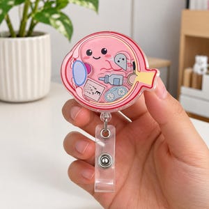 May include: A circular acrylic badge reel featuring a cartoon eye illustration with a smiling face. The design includes small icons of an eye chart with the letter E, eye drops, contact lenses, and a magnifying glass. The badge reel has a clear plastic strap.