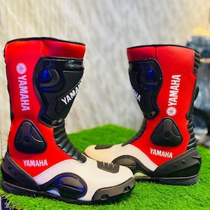 Puede incluir: Un par de botas de moto de caña alta en rojo, blanco y negro. Las botas presentan el logotipo y el texto de Yamaha impresos en blanco sobre las secciones rojas. Incluyen paneles protectores negros moldeados en tobillos, espinillas y punteras.