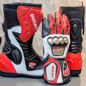 Puede incluir: Un par de botas de moto Ducati Corse en negro, blanco y rojo junto a un guante a juego. Las botas presentan espinilleras rojas con el logotipo de Ducati. El guante incluye un protector metálico para los nudillos y el texto SCP CONCEPT.