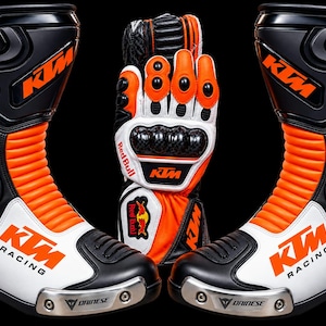 Puede incluir: Un par de botas de carreras KTM negras, blancas y naranjas con deslizadores metálicos en la punta y un guante de motocicleta a juego con la marca Red Bull, protección de carbono en los nudillos y el logotipo de KTM sobre fondo negro.