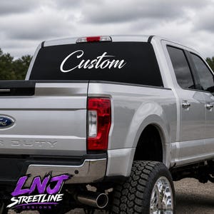 May include: A silver pickup truck with a white vinyl decal applied to the rear window. The decal features the word Custom written in a white cursive script font. The truck has a chrome bumper, red taillights, and large off-road tires.