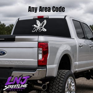 May include: A white vinyl decal applied to the rear window of a silver pickup truck. The decal features a flying duck silhouette positioned above the numbers 570. The text Any Area Code is displayed above the truck in the image background.