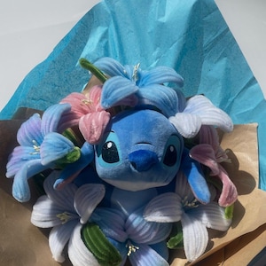 May include: A blue plush Stitch toy surrounded by blue, pink, and white fabric flowers. The bouquet is wrapped in brown paper and blue tissue paper, creating a colorful and whimsical arrangement.