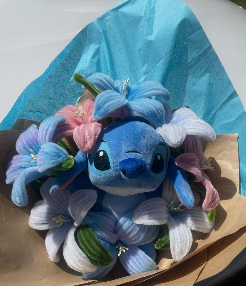 May include: A blue plush Stitch toy surrounded by blue, pink, and white fabric flowers. The bouquet is wrapped in brown paper and blue tissue paper, creating a colorful and whimsical arrangement.