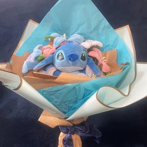 May include: A plush blue Stitch character bouquet with pink and blue flowers. The bouquet is wrapped in brown and blue paper, tied with a blue ribbon, and presented on a dark blue surface.