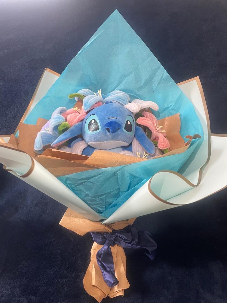May include: A plush blue Stitch character bouquet with pink and blue flowers. The bouquet is wrapped in brown and blue paper, tied with a blue ribbon, and presented on a dark blue surface.