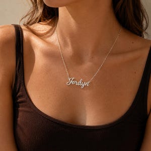 May include: A silver Jordyn name necklace. The delicate cursive pendant hangs from a fine chain. The necklace is displayed against a dark brown vest top.