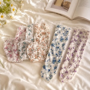 Puede incluir: Una colección de calcetines de encaje blanco transparente con delicados patrones florales en varios colores, incluyendo rosa, naranja, azul y morado. Los calcetines están dispuestos en grupo, algunos doblados y otros extendidos.