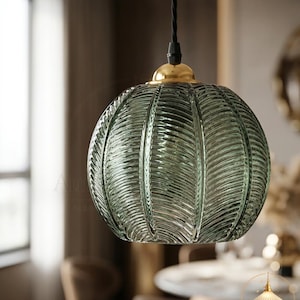 May include: A spherical pendant light featuring translucent sage green glass with a textured, ribbed leaf-like pattern. The glass shade is topped with a polished gold-tone metal cap and suspended by a black twisted fabric cord.