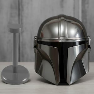 Puede incluir: Un casco plateado de Mandaloriano con visera negra, junto a un soporte gris para el casco. El casco presenta un dise&ntilde;o detallado con crestas verticales en la parte superior. El soporte tiene un dise&ntilde;o sencillo en forma de T.