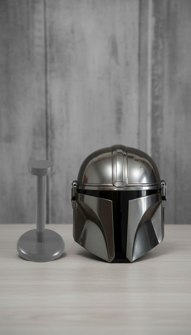 Puede incluir: Un casco plateado de Mandaloriano con visera negra, junto a un soporte gris para el casco. El casco presenta un dise&ntilde;o detallado con crestas verticales en la parte superior. El soporte tiene un dise&ntilde;o sencillo en forma de T.