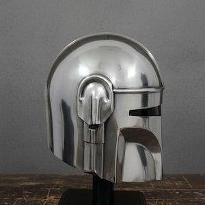 Puede incluir: Un casco plateado pulido con una visera y orejeras distintivas, exhibido sobre un soporte negro. El dise&ntilde;o del casco presenta una parte superior redondeada y una visera en forma de T. El casco es una r&eacute;plica de un personaje de ciencia ficci&oacute;n popular.