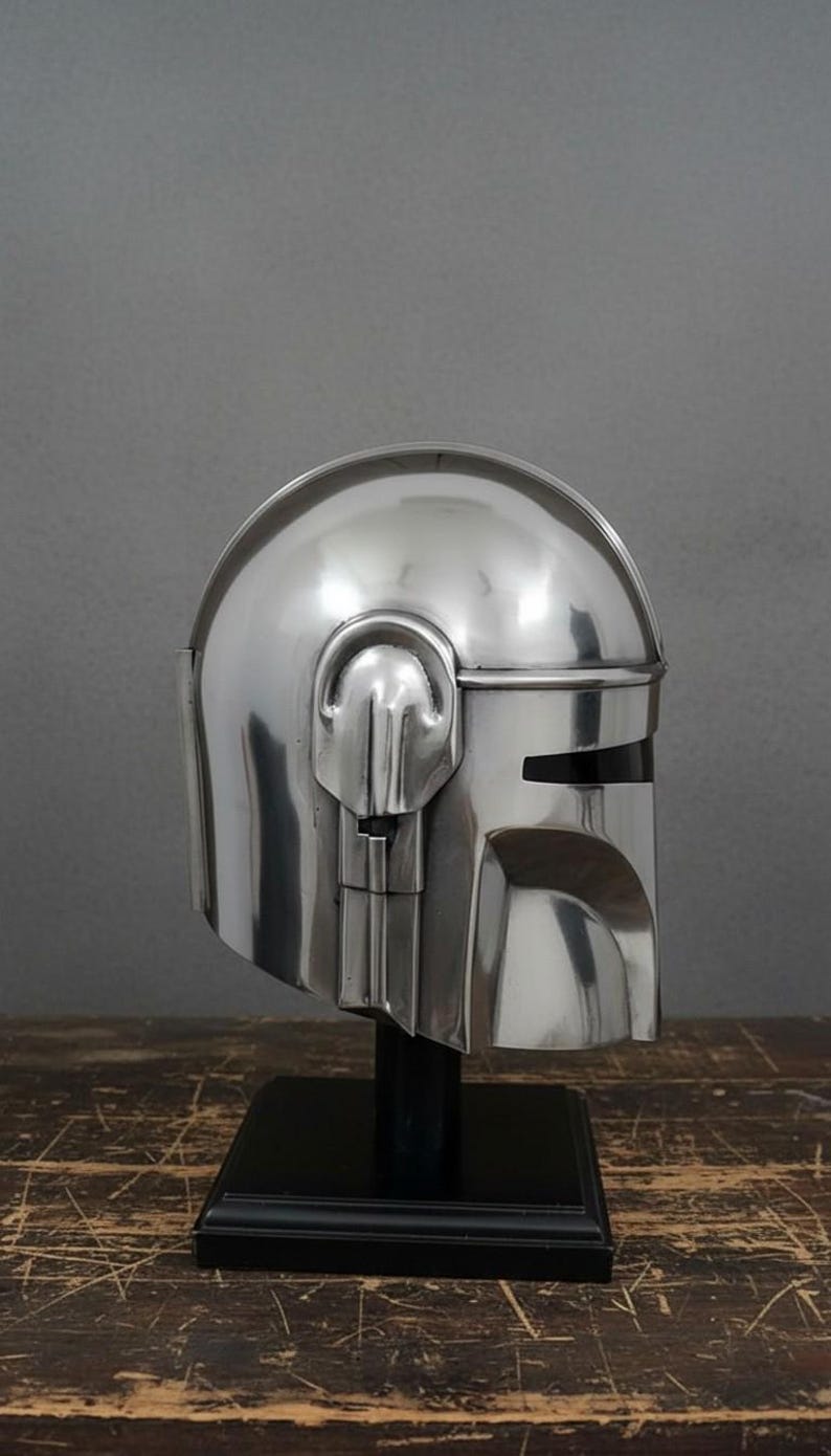 Puede incluir: Un casco plateado pulido con una visera y orejeras distintivas, exhibido sobre un soporte negro. El dise&ntilde;o del casco presenta una parte superior redondeada y una visera en forma de T. El casco es una r&eacute;plica de un personaje de ciencia ficci&oacute;n popular.