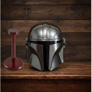 Puede incluir: Un casco plateado de Mandaloriano con visera negra sobre una superficie de madera. A la izquierda, un soporte para casco granate. El casco tiene un dise&ntilde;o detallado y es una r&eacute;plica de la serie Star Wars.