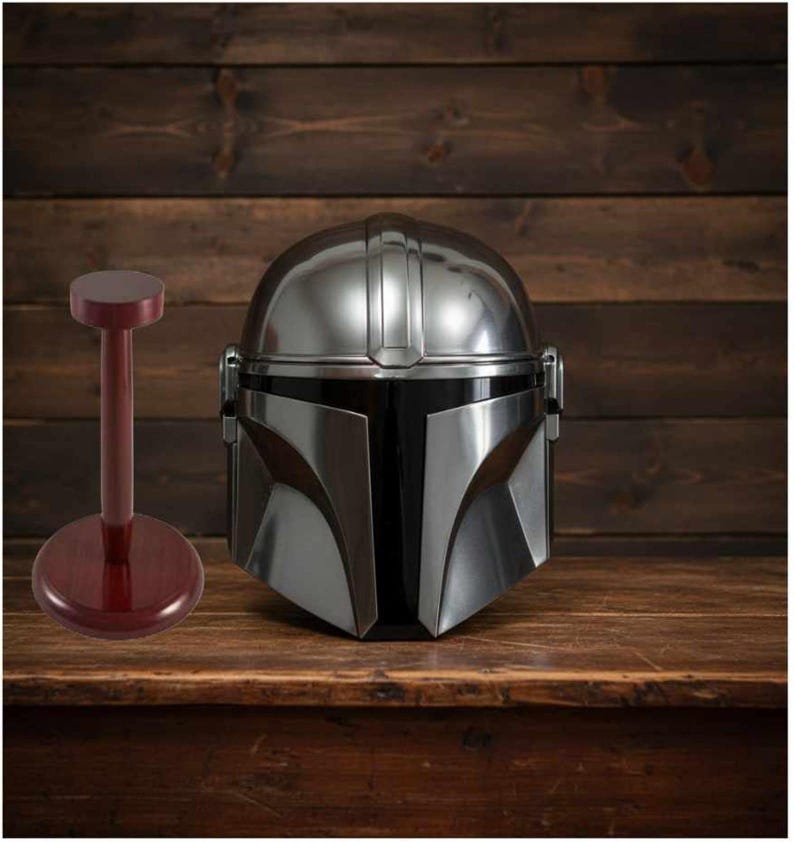 Puede incluir: Un casco plateado de Mandaloriano con visera negra sobre una superficie de madera. A la izquierda, un soporte para casco granate. El casco tiene un dise&ntilde;o detallado y es una r&eacute;plica de la serie Star Wars.